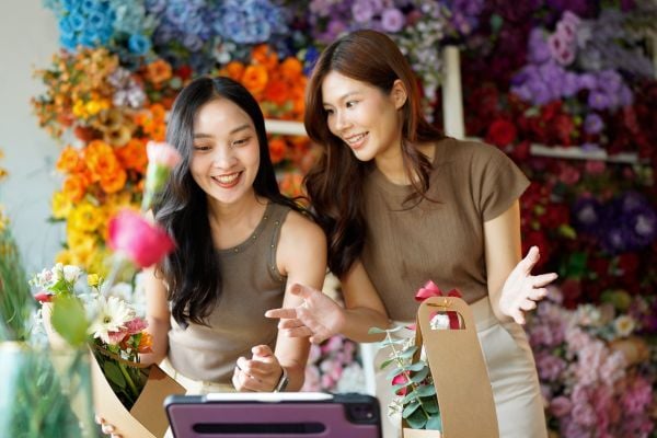 Two woman in front of a laptop with flowers behind them and a promotional item kit on the table before them.